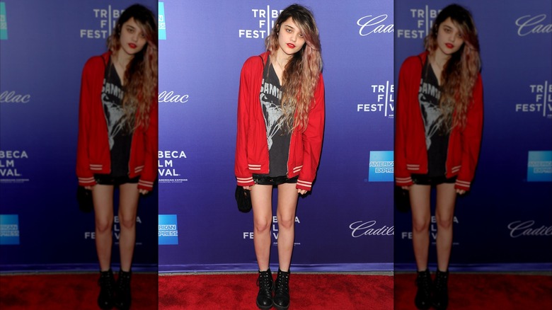 Sky Ferreira posant lors d'un événement sur le tapis rouge, la tête sur le côté, vêtue d'une veste rouge, d'une chemise de groupe, d'un short noir et de bottes de combat.