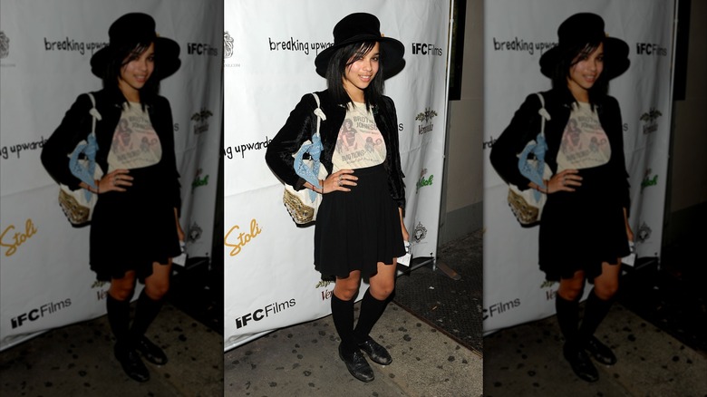 Zoë Kravitz posant lors d'une première de film avec la main sur les hanches, portant une veste et une jupe noires avec un t-shirt graphique blanc et un chapeau noir à larges bords.