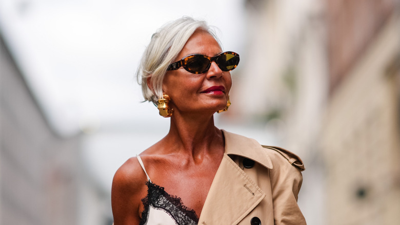 Une femme pose avec des lunettes de soleil ovales et un trench-coat beige pend sur son épaule