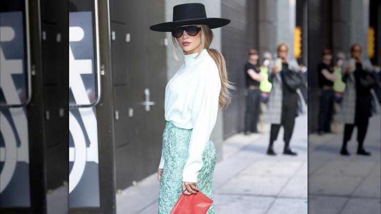Jennifer Lopez posant dehors à New York portant un chapeau noir à larges bords avec des lunettes de soleil, un haut blanc et une jupe verte.