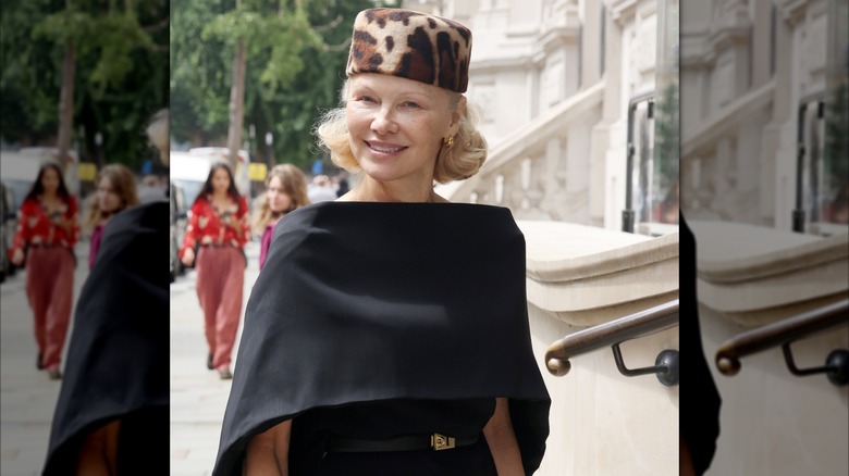 Pamela Anderson souriante dehors, vêtue d'une robe caplet noire avec un chapeau casemate à imprimé léopard.