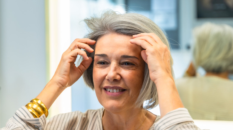 Femme mûre souriant et fixant ses cheveux avec un miroir derrière elle.