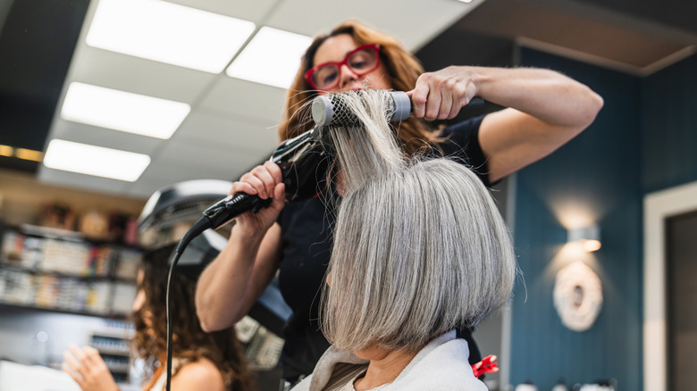 Femme avec un carré gris dans un salon se faisant coiffer avec un sèche-cheveux et une brosse.