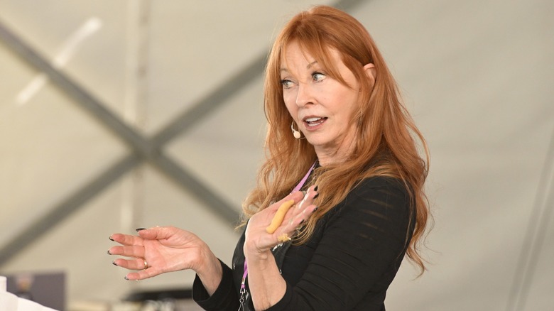 Cassandra Peterson donne une conférence vêtue d'un haut noir avec ses longs cheveux roux ondulés détachés.