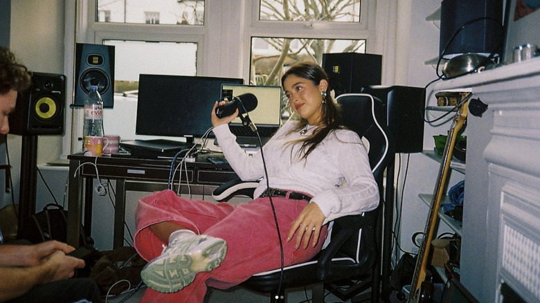 Lola Consuelos portant un pantalon rose vif et un haut blanc, assise sur une chaise de jeu tenant un micro d'enregistrement.