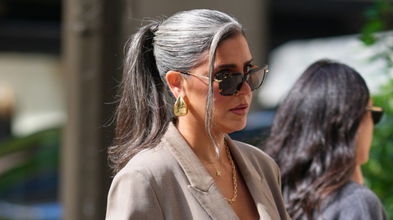 Une femme aux cheveux gris en queue de cheval haute portant des lunettes de soleil, des bijoux en or et un blazer gris.