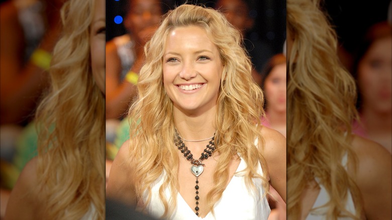 Kate Hudson dans un gilet court blanc avec un gros collier noir et ses cheveux en vagues lâches avec un pouf bang.