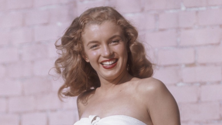 Marilyn Monroe posant dans un haut blanc avec du rouge à lèvres et des cheveux ondulés blond fraise devant un mur de briques.