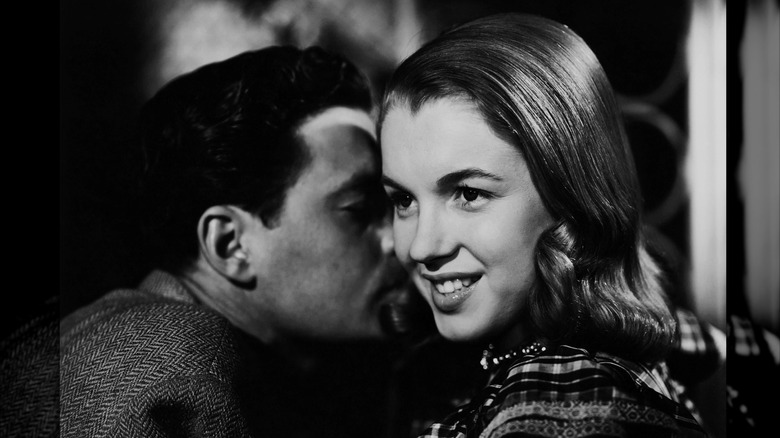 Photo en noir et blanc de Marilyn Monroe avec des cheveux bruns élégants, souriant tandis qu'un homme lui chuchote à l'oreille.