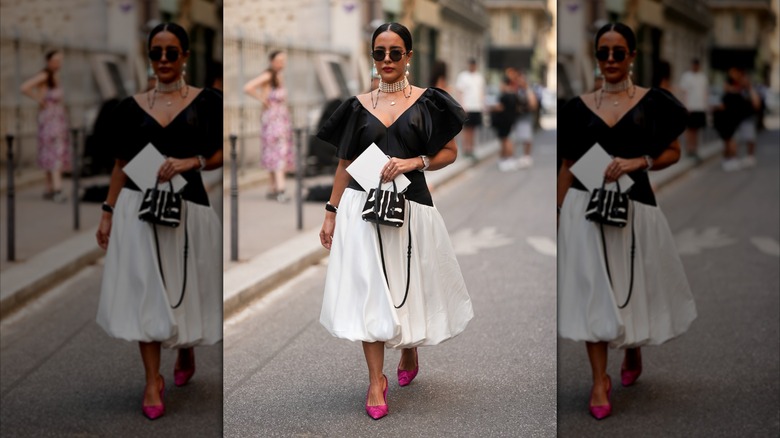 Une femme portant une robe noire et blanche, un tour de cou à plusieurs rangs de perles, des lunettes de soleil noires et des talons rose vif.