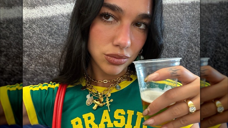 Selfie de Dua Lipa portant un collier à breloques et une bague tendance alors qu'elle tient un verre de bière