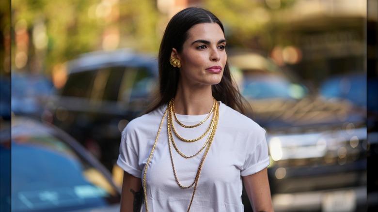 Une femme portant un t-shirt blanc à manches retroussées et plusieurs colliers en chaîne dorée.