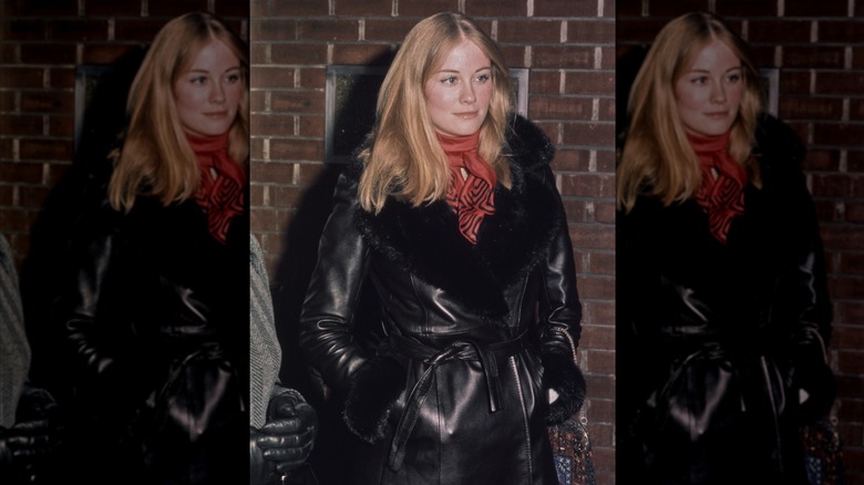 Cybill Shepherd, les mains dans les poches, portant un manteau en cuir Penny Lane et un foulard rouge.