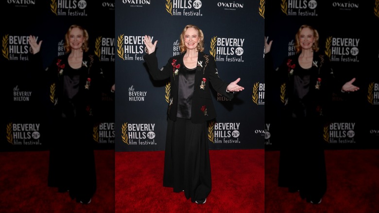 Cybill Shepherd sourit à bras ouverts portant un blazer noir fleuri lors d'un événement sur le tapis rouge.