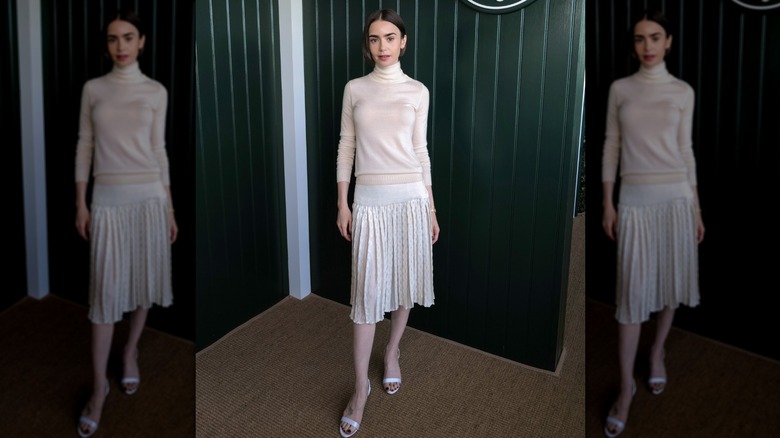 Lily Collins porte un haut à col roulé crème et une jupe plissée blanche avec des sandales blanches lors d'un événement.