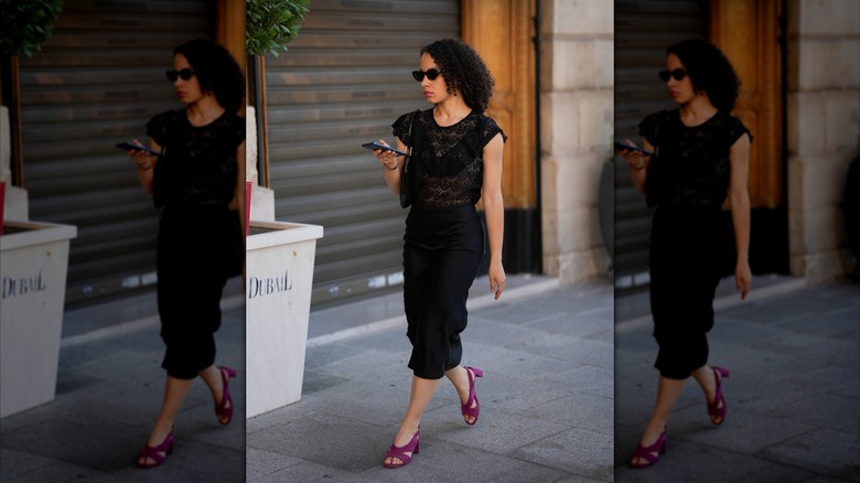 Une femme marchant dans la rue portant une jupe crayon noire, un haut noir en dentelle et des talons carrés violets.