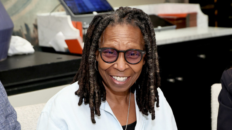 Whoopi Goldberg souriant lors d'un événement vêtu d'une chemise blanche avec des lunettes teintées noires.