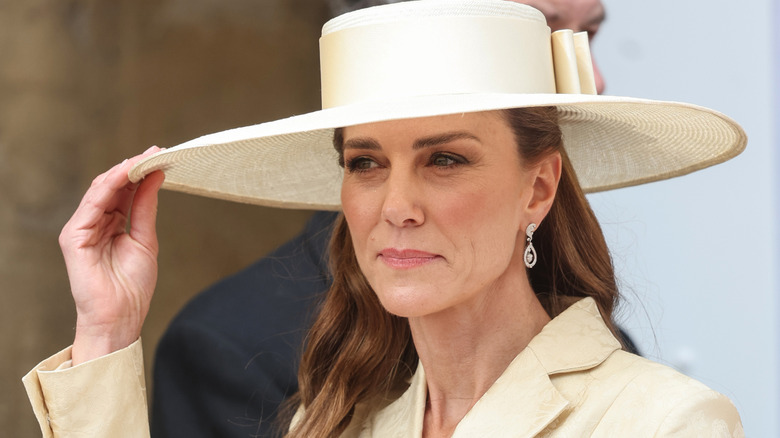 Kate Middleton touchant le bord de son chapeau jaune portant un blazer assorti avec des boucles d'oreilles en diamant.