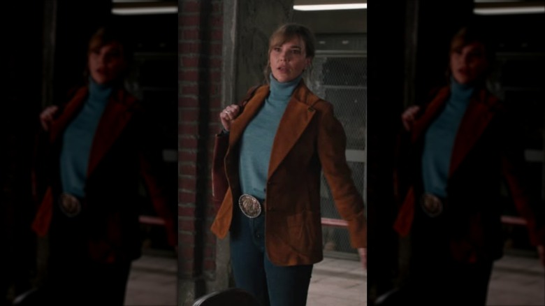 Belle Skinner portant un col roulé turquoise et un blazer en daim marron