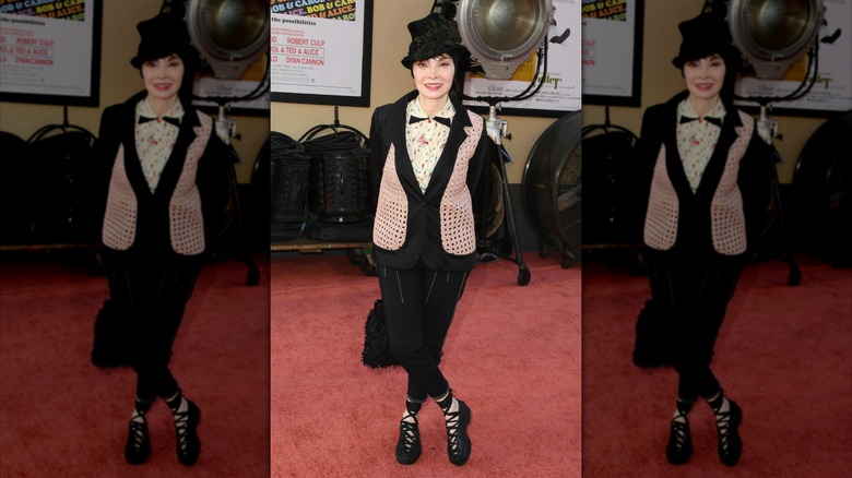 Toni Basil posant sur le tapis rouge avec les chevilles croisées, vêtue d'une tenue noire aux accents roses et blancs.