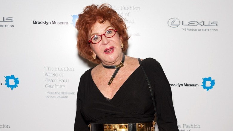 Sally Jessy Raphael portait un haut noir décolleté avec une ceinture dorée brillante et un collier de ceinture assorti ainsi que ses cheveux roux courts et ses lunettes rouges emblématiques lors de sa participation au Fashion World de Jean Paul Gaultier à New York en 2013.