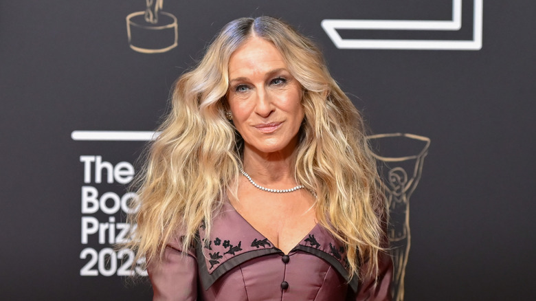 Sarah Jessica Parker posant lors d'un événement vêtue d'une robe marron aux cheveux ondulés et d'un collier délicat.