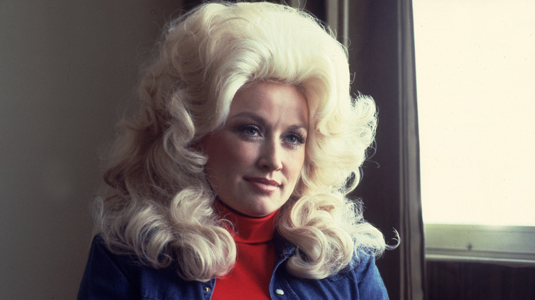 Dolly Parton porte une perruque blonde volumineuse, un col roulé rouge et une chemise bleue