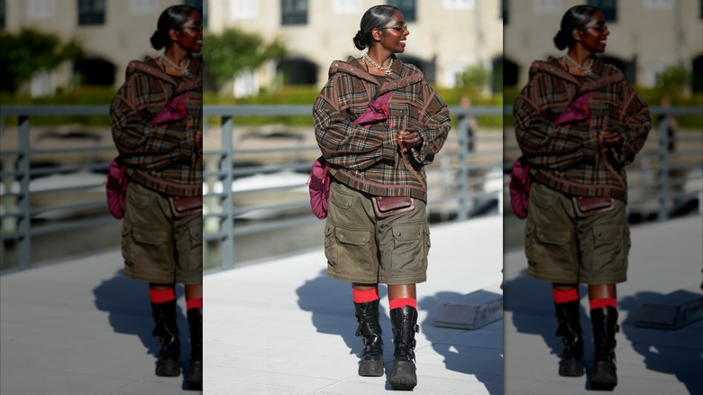Une femme portant une veste tartan marron et olive, un short cargo olive et des bottes noires.