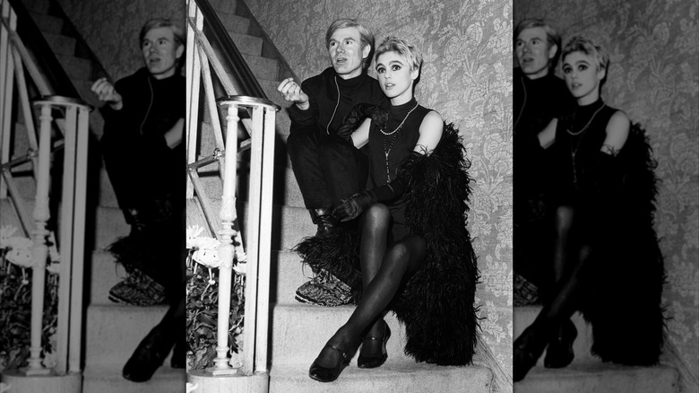 L'artiste Andy Warhol vêtu de noir et Edie Sedgwick portant une tenue noire avec un boa en plumes noires, assis sur un escalier.