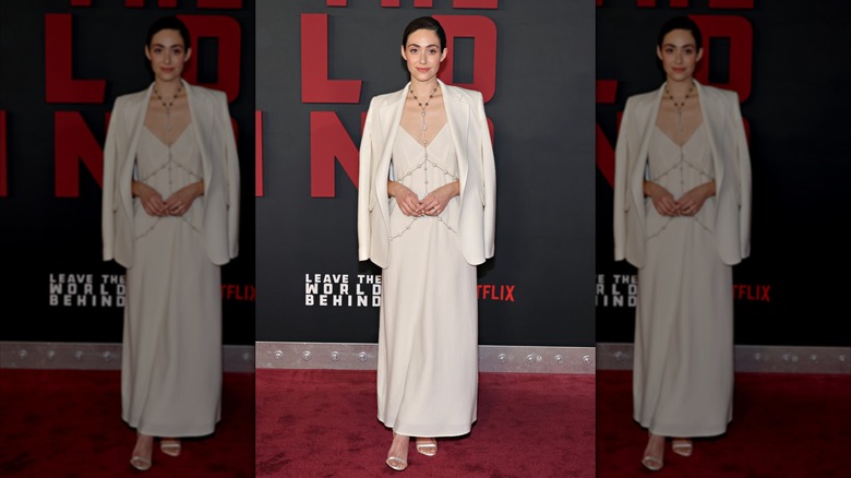 Emmy Rossum porte un blazer blanc et une robe avec une longue chaîne de corps argentée