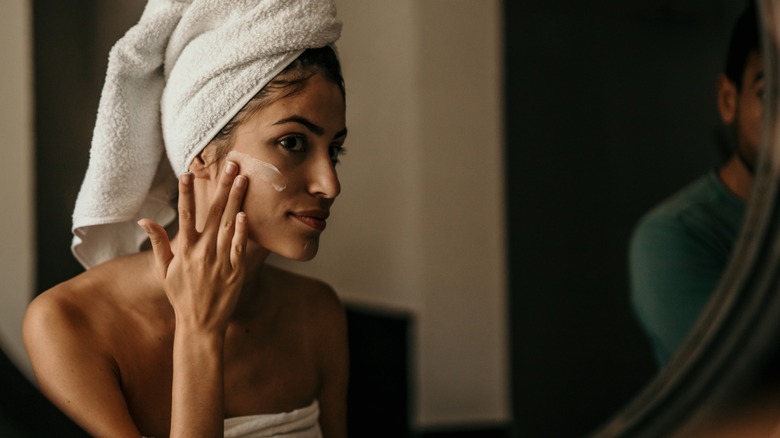 Une femme vêtue d’une serviette applique une lotion sur son visage.