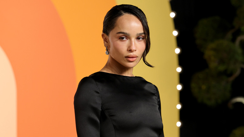 Zoë Kravitz pose devant un fond orange et jaune tout en portant une robe noire et un chignon.