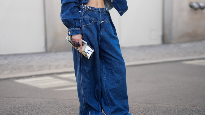 Une femme porte un jean baggy taille basse et des tongs tout en tenant un magazine Vogue, son téléphone et ses lunettes de soleil