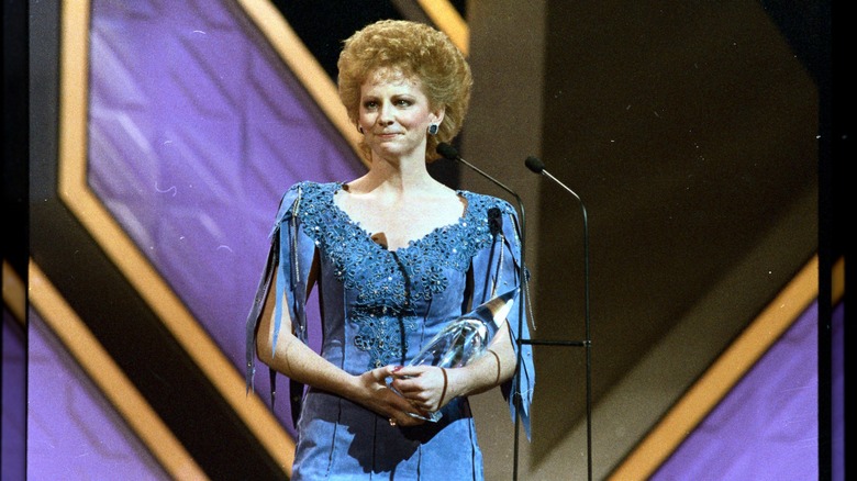 Reba McEntire avec une coiffure courte et volumineuse portant une robe bleue sur scène lors des Country Music Association Awards en 1986.