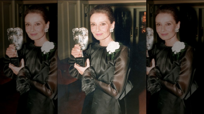Audrey Hepburn posant avec une récompense, vêtue d'une robe noire à manches transparentes et ornée d'une rose blanche.