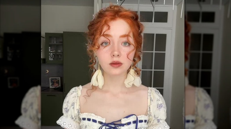 Une rousse avec un maquillage cottagecore et des boucles d'oreilles fleuries posant pour une photo