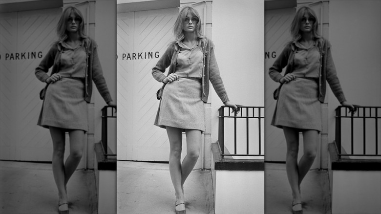 Jean Shrimpton pose avec la main sur la hanche, portant une jupe courte et de grosses lunettes de soleil