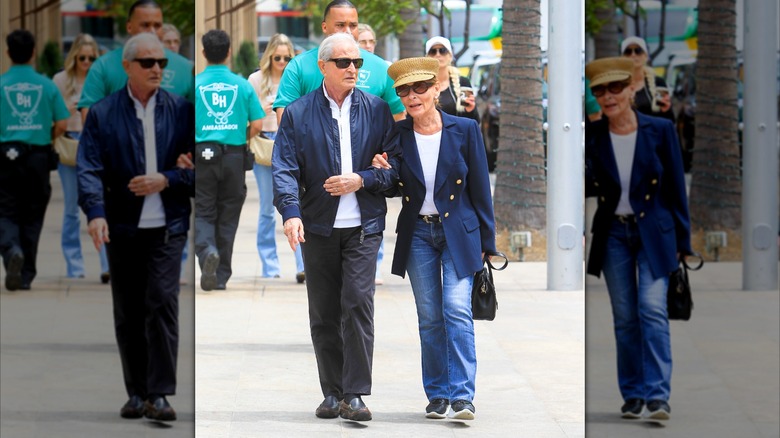 Judy Sheindlin marche avec son mari (à gauche) portant un jean évasé, une chemise à manches longues blanche, un blazer bleu et un chapeau de pêcheur.