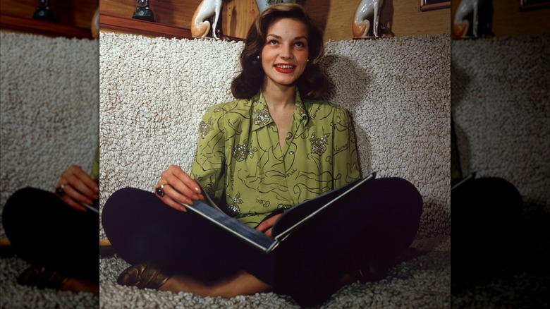 Lauren Bacall assise les jambes croisées avec un livre ouvert sur ses genoux, vêtue d'un chemisier vert orné et d'un rouge à lèvres rouge vers 1945