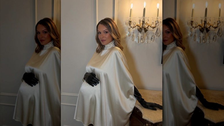 Kayla Wallace porte une robe à col roulé en soie blanche et des gants en cuir noir, la main sur son baby bump.