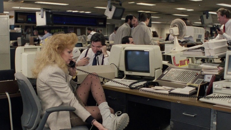 Melanie Griffith assise à un bureau portant un blazer beige, une jupe à carreaux, des collants à pois et des baskets blanches. 