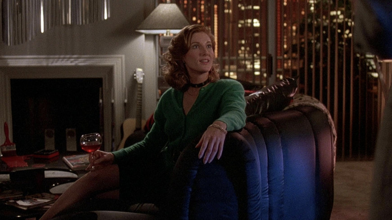 Annette O'Toole assise sur un canapé, tenant un verre de vin, vêtue d'une chemise verte avec un tour de cou noir. "Traverse mon cœur."